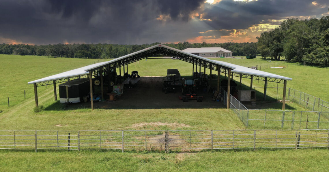 Open-sided agricultural pole barn for RV storage, farm equipment, and livestock shelter with fenced area on a farm property.